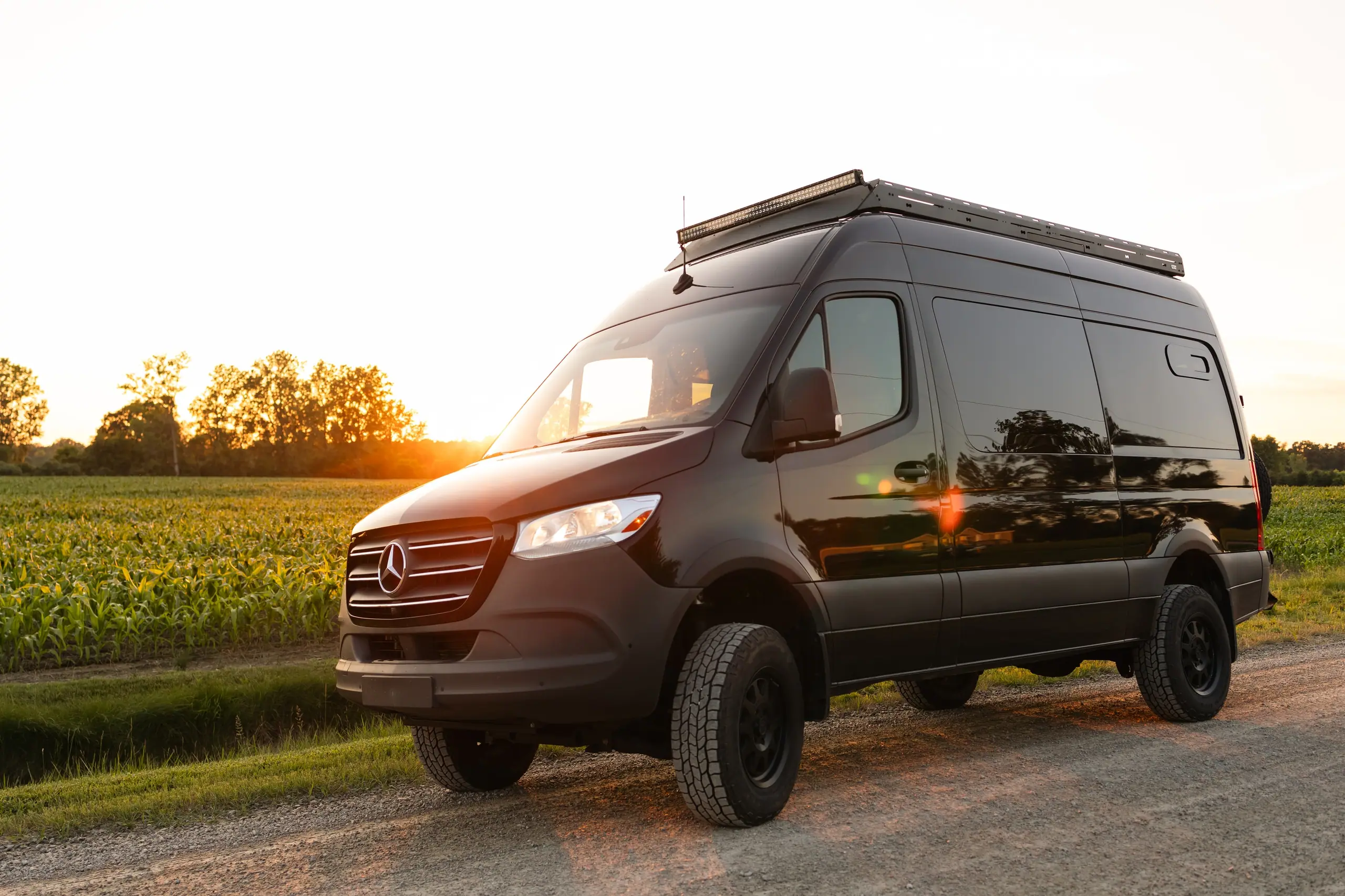 Black Mercedes-Benz Sprinter camper van with a roof rack, parked on a gravel road beside a cornfield at sunset