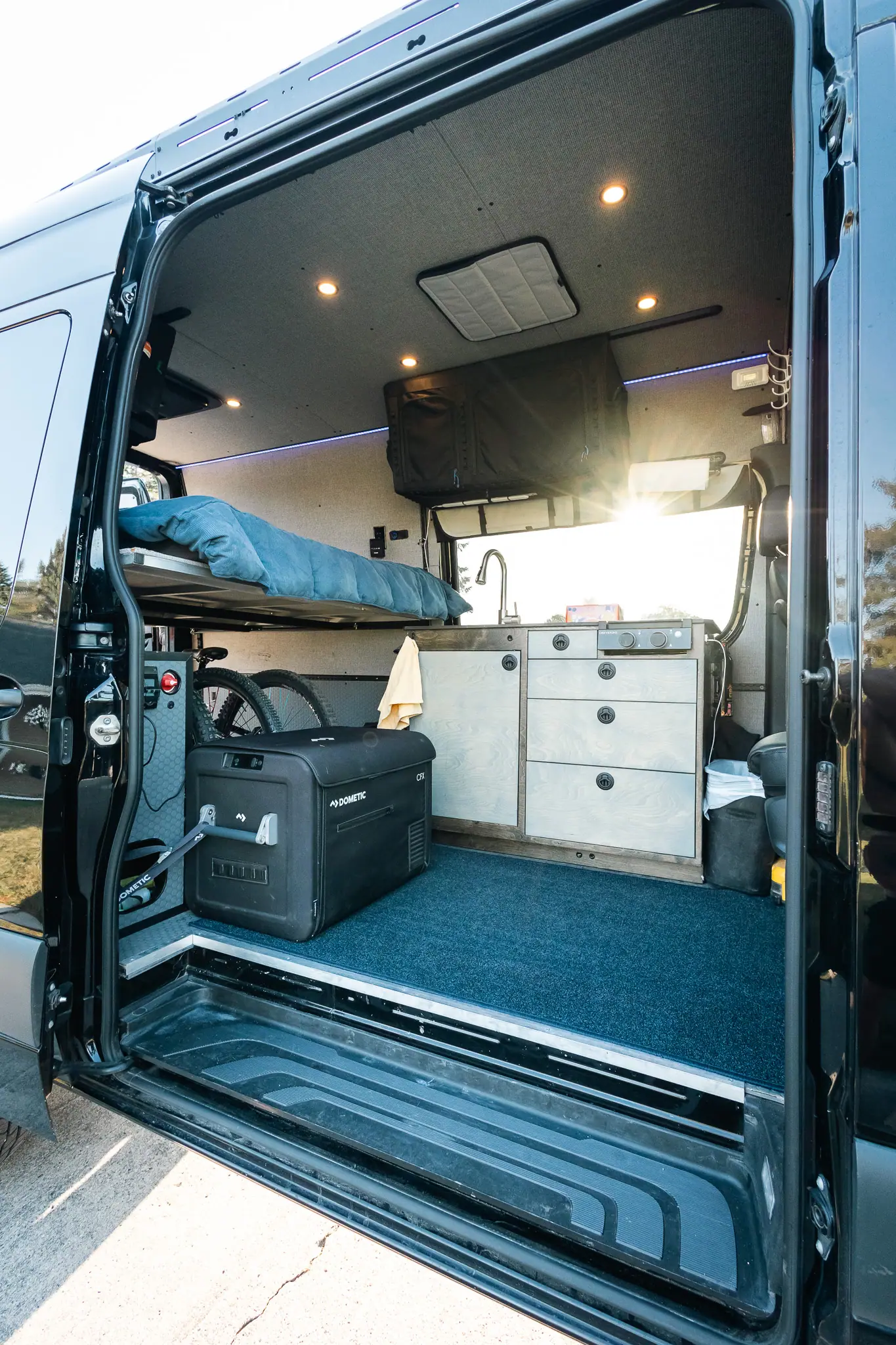 View through the sliding door of a Sprinter camper van: galley, fridge, raised rear bed and bike storage
