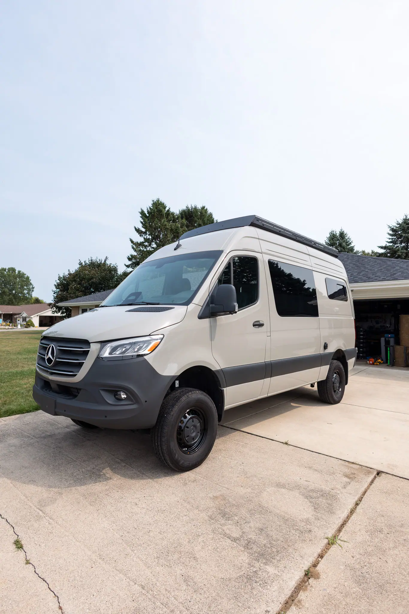 Sand-gray Mercedes-Benz Sprinter high-roof camper van with a black roof rack and black steel wheels