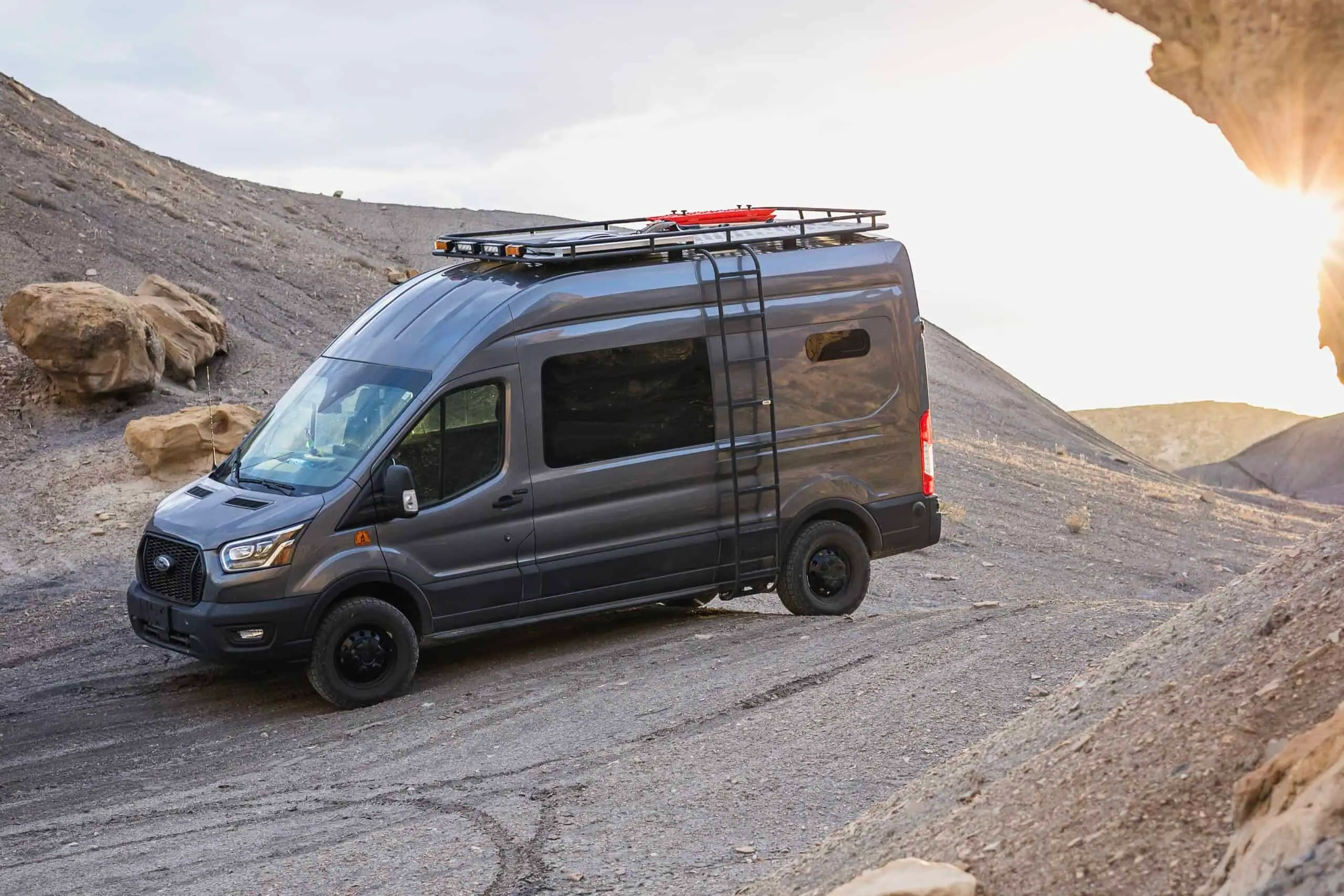 Dark gray Ford Transit high-roof camper van with a roof rack, ladder and all-terrain tires on a dirt road
