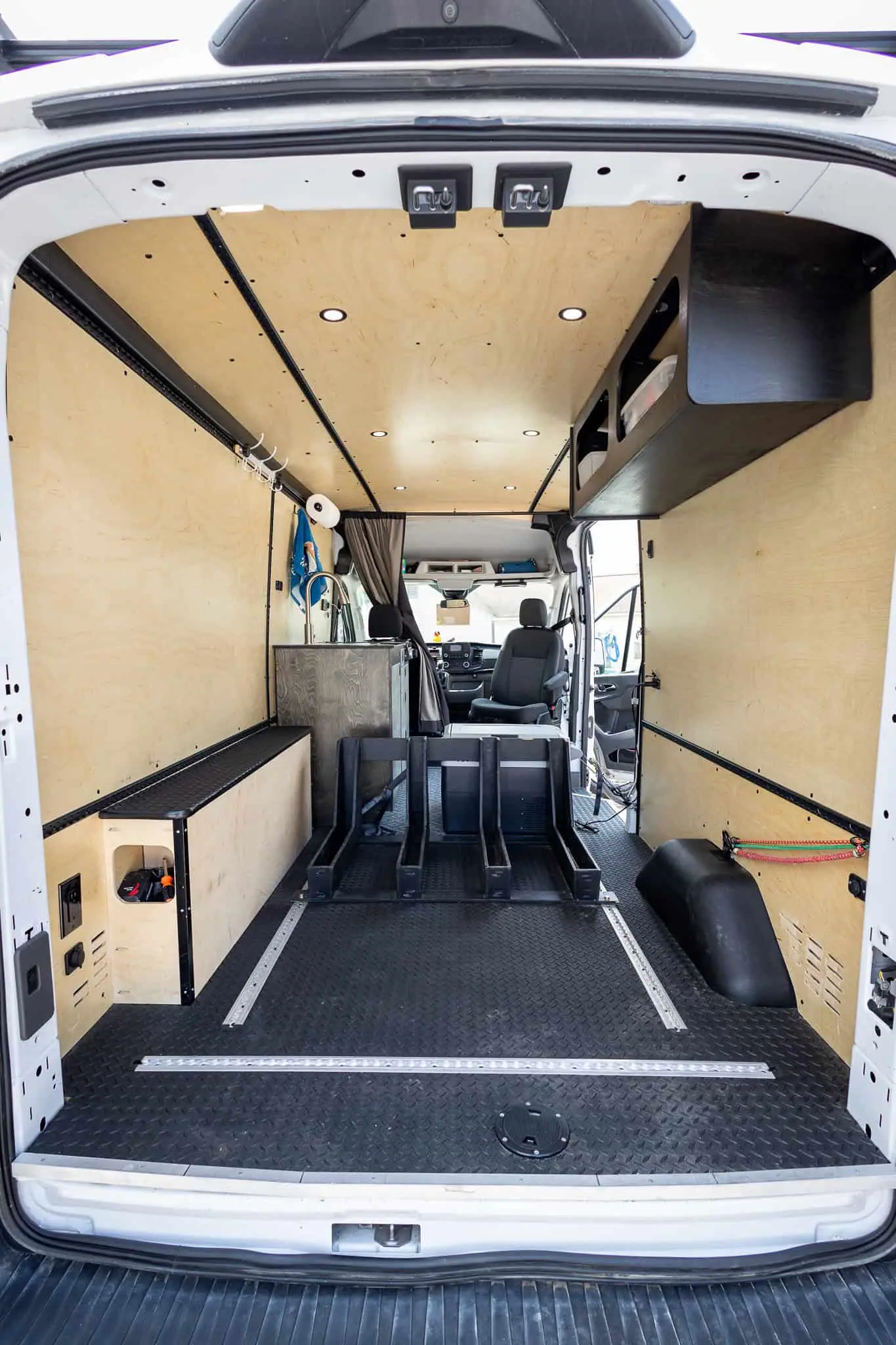 Open cargo floor with L-track rails and plywood walls, seen through the rear doors of a Ford Transit van
