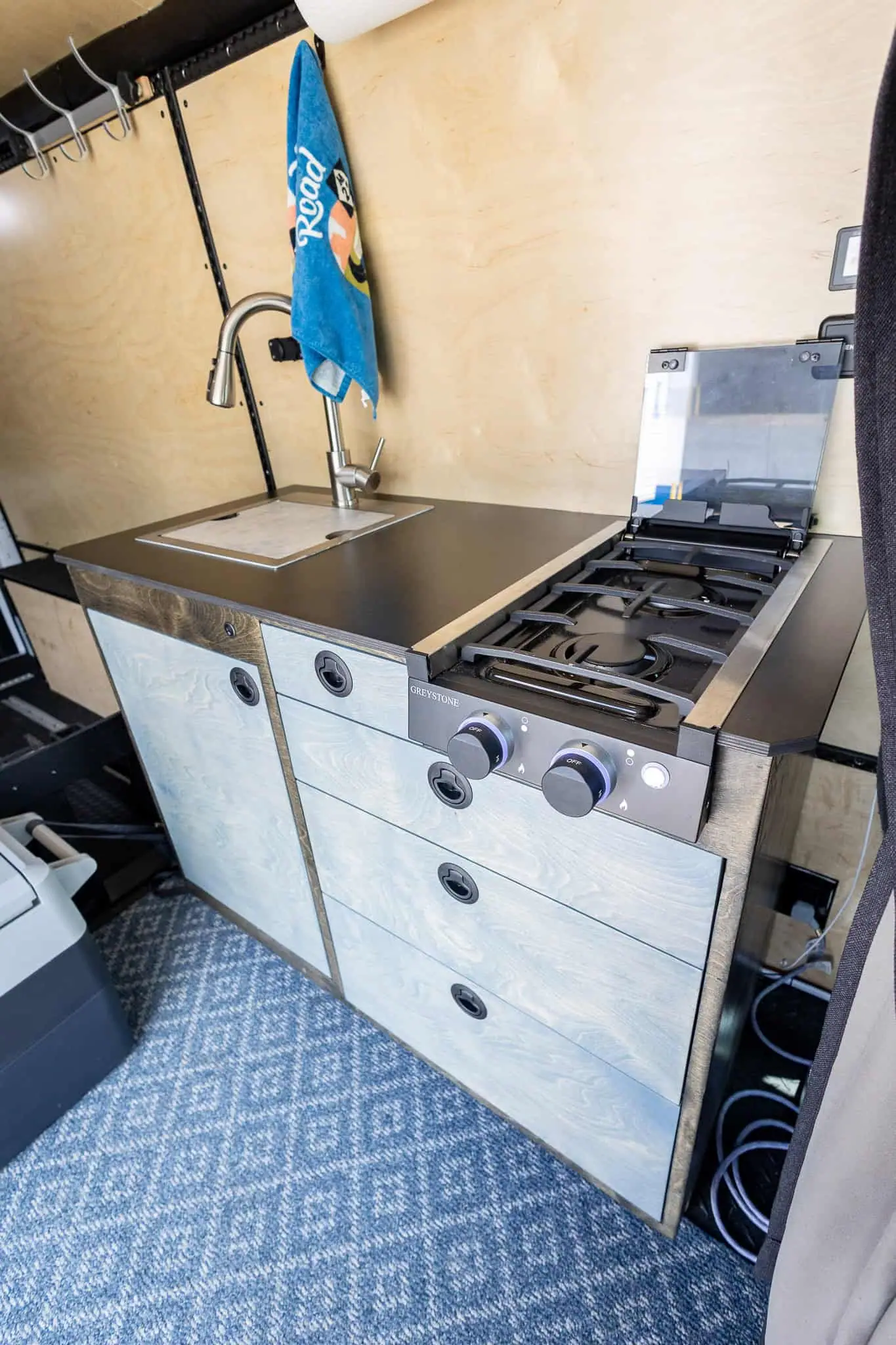 Compact van galley with blue-gray cabinets, a stainless sink and a two-burner stove